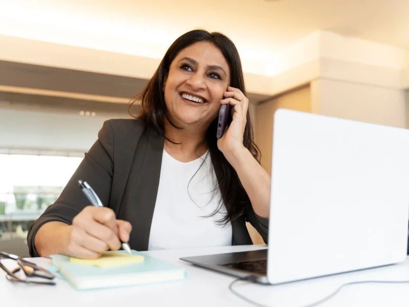 Happy professional woman sitting in front of laptop using phone to make NDIS referral